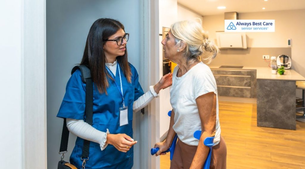 A caregiver and a senior woman in a home setting, with the caregiver providing assistance and offering gentle support. The image captures a moment of trust and care between the senior and her caregiver.